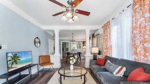A living room with a ceiling fan, a television, a sofa, a coffee table, and a dining area in the background. - Cozy Springfield Abode - AC, Wifi, WD (Jacksonville)