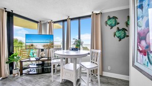 Una habitación con una mesa redonda y cuatro sillas, un ventanal con cortinas, un cuadro de una playa y una planta en maceta.