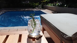 Une table en bois sur laquelle se trouve un récipient en verre rempli de glace et une branche de thym, un fauteuil de relaxation blanc avec coussin, et une piscine en arrière-plan.