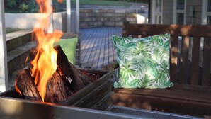 Un banc en bois avec un coussin à motifs verts, un foyer avec des bûches qui brûlent et un mur de pierre en arrière-plan.