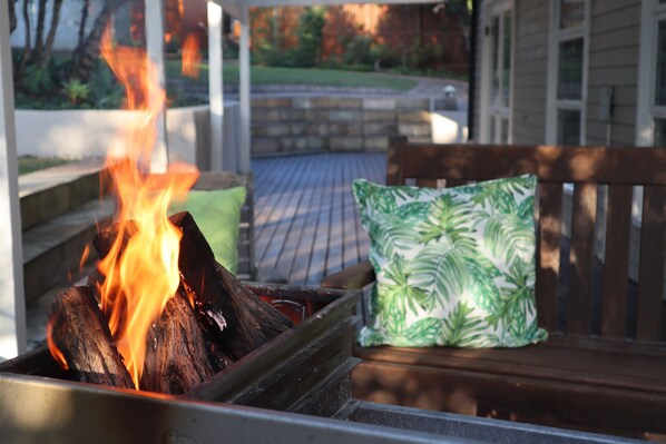A wooden bench with a green patterned cushion, a fire pit with burning logs, and a stone wall in the background.