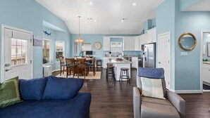 A spacious living area with a blue couch, a dining table, and a kitchen in the background. - AJs Beach Haven - family fun- beautiful vacation home in Sea Isle (Galveston)