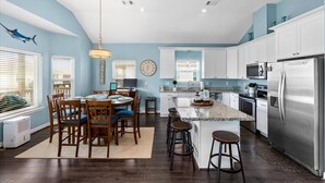A modern kitchen with white cabinets, a stainless steel refrigerator, and a dining area with wooden chairs and a table. - AJs Beach Haven - family fun- beautiful vacation home in Sea Isle (Galveston)