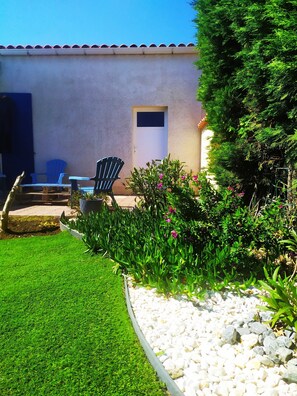 Un espace patio avec un mur blanc, une porte et une petite fenêtre. On y trouve deux chaises bleues et un pot de fleurs avec des fleurs roses.