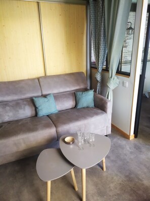 Un salon moderne avec un canapé gris, deux coussins à motifs, une petite table ronde sur laquelle se trouve un bol et quatre verres à vin, et une fenêtre avec un rideau.
