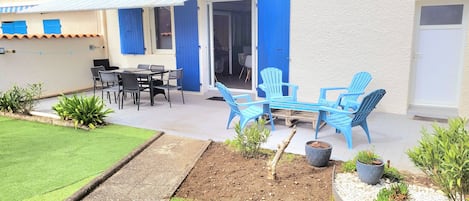 A patio area with a table and chairs, a small garden, and a building with blue shutters and a white exterior.