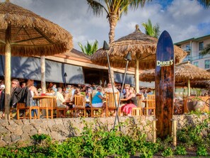 An outdoor dining area with thatched umbrellas, wooden chairs, and a stone wall. - HONUA KAI RESORT-Hokulani 314 - 106 ⭐️⭐️⭐️⭐️⭐️ FIVE STAR REVIEWS - OCEAN & MNT VIEWS (Lahaina)