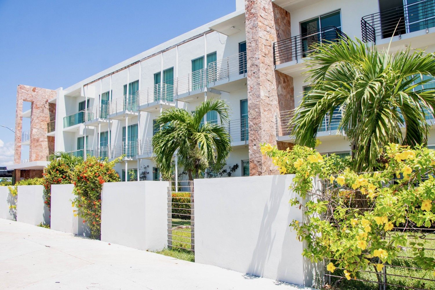 A modern multi-story building with balconies, a white facade, and surrounding greenery.