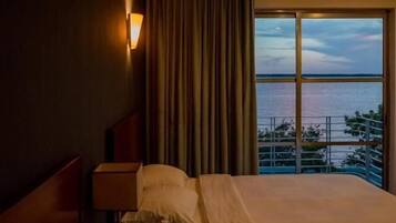 Un cuarto de hotel con una cama, una mesita de noche, una lámpara y vistas al mar a través de amplias ventanas.