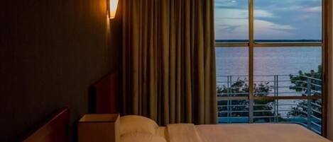 A hotel room with a bed, a nightstand, a lamp, and a view of the sea through large windows.