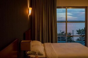 Un cuarto de hotel con una cama, una mesita de noche, una lámpara y vistas al mar a través de amplias ventanas.