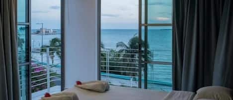 Quarto de hotel com uma cama grande, duas janelas com vista para o mar e um horizonte urbano distante.