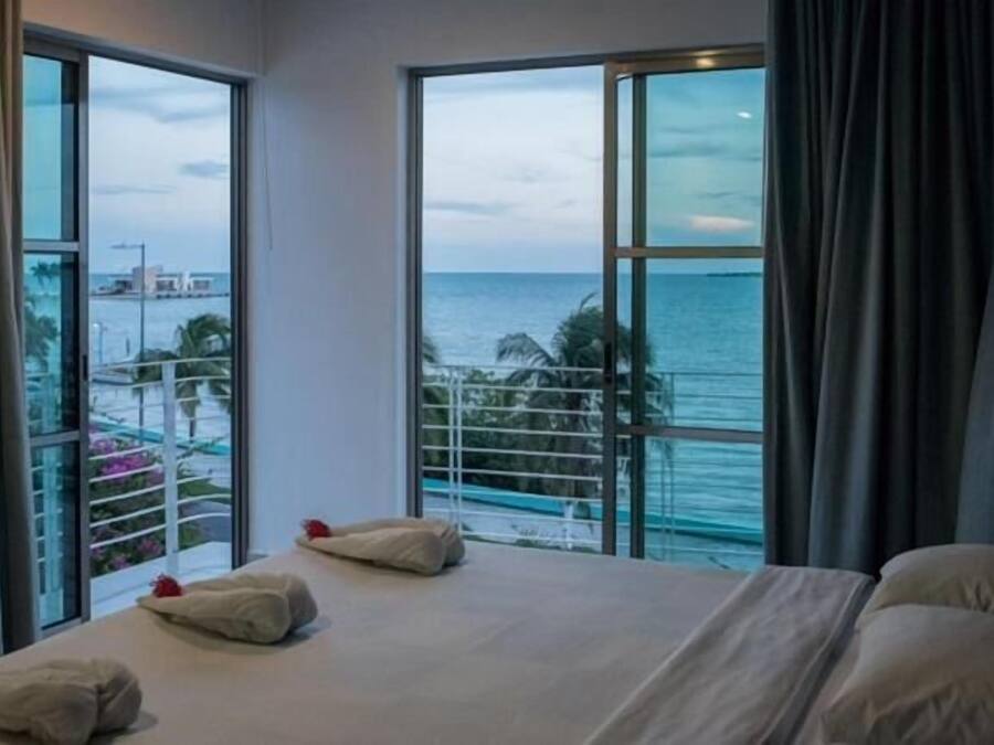 Quarto de hotel com uma cama grande, duas janelas com vista para o mar e um horizonte urbano distante.