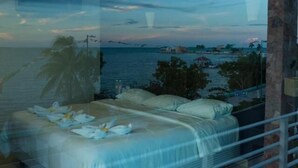 A bedroom with a bed, pillows, and a view of the sea and distant buildings. - Hotel Noor (Chetumal)