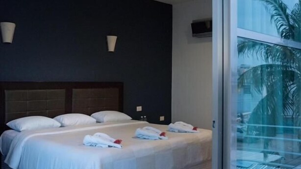 A hotel room with a large bed, two bedside tables, a TV mounted on the wall, and a window with a view of palm trees. - Hotel Noor (Chetumal)