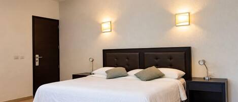 Quarto de hotel com cama, duas mesinhas de cabeceira e duas luzes fixadas na parede.