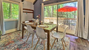A dining area with a wooden table set for four, white chairs, and a view of a deck with outdoor furniture and a red umbrella. - Pet-Friendly Stanwood Sanctuary w/ Fire Pit! (Stanwood)