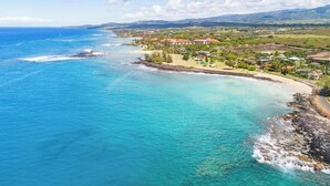 An aerial view of a coastal area with clear turquoise waters, a sandy beach, and a residential area with houses and greenery. - Waikomo Stream Villas 332 – 2 Bedroom Loft, A/C, PBAC Access (Koloa)