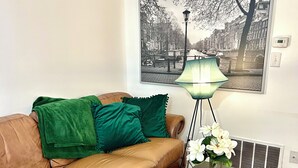 A living room with a brown leather sofa, green cushions, a black coffee table with a magazine, a floor lamp, and a framed black and white cityscape picture on the wall.