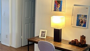 A small, neatly arranged office space with a wooden desk, a single chair, a lamp, and framed artwork on the wall.