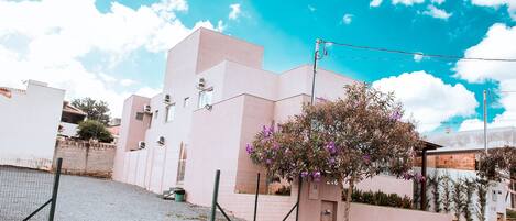 Un edificio moderno con fachada rosa, un pequeño árbol con flores moradas y un cielo azul despejado.