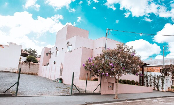 Un bâtiment moderne à la façade rose, un petit arbre aux fleurs violettes et un ciel bleu clair.