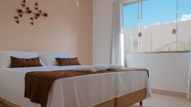 A bedroom with a bed, white walls, a window with curtains, and a decorative wall piece. - Pousada Shalom Capitólio (Capitólio)