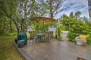 A wooden deck with a table and chairs under a large umbrella, surrounded by greenery and trees. - Modern Home ~ 2 Mi to Cranmore Mountain Resort! (Conway)