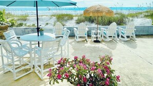 A beachside patio with white chairs and a table, a blue umbrella, and a thatched structure.