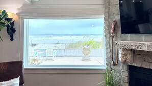 A living room with a large window overlooking the ocean, a stone fireplace, and a flat-screen TV mounted on the wall.