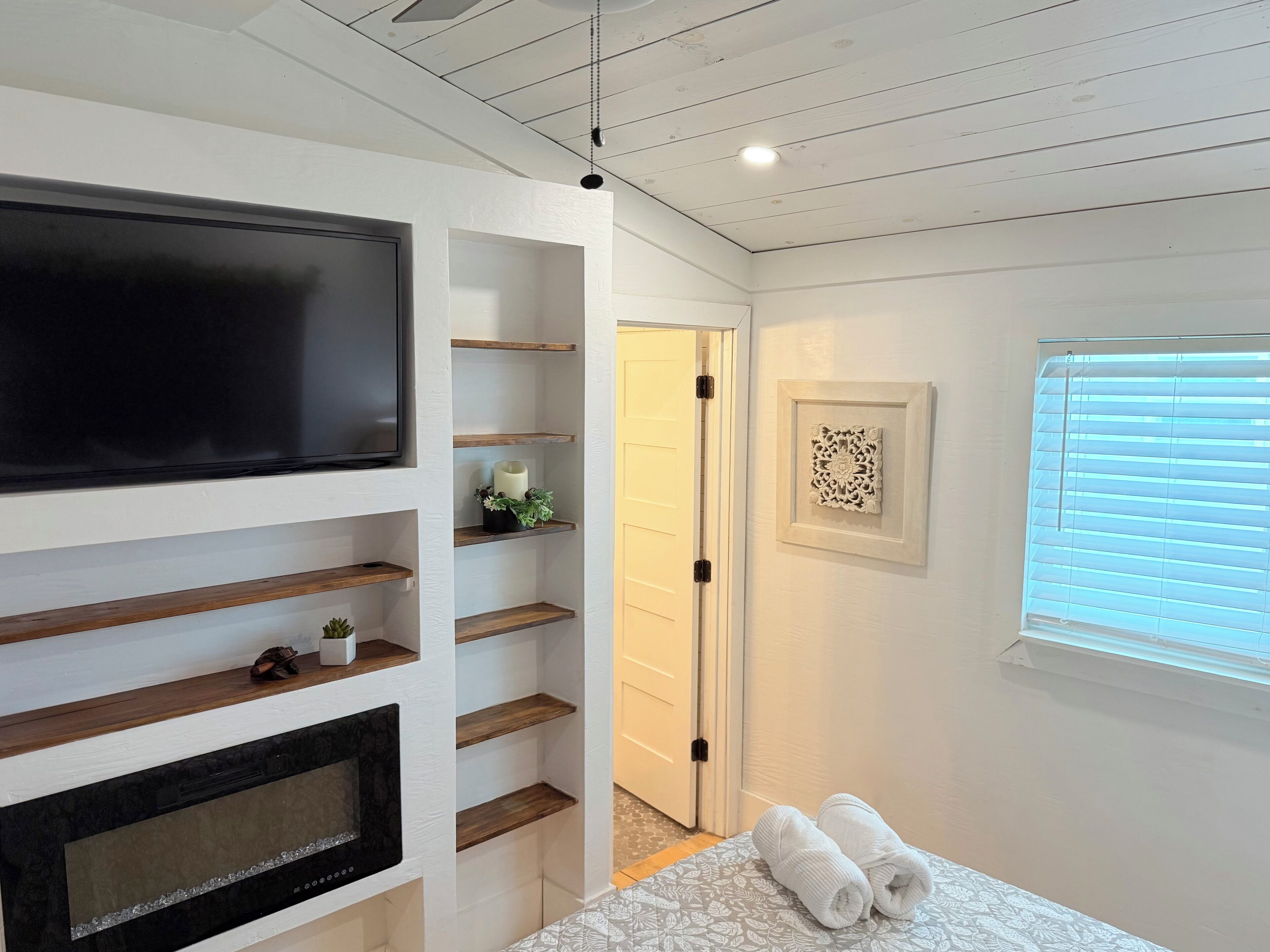 A modern bedroom with a flat-screen TV, wooden shelves, a fireplace, and a bed with rolled towels.