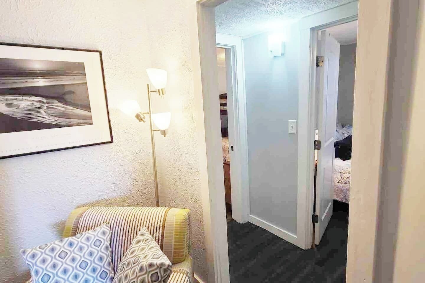 Une chambre d’hôtel avec un lit, une chaise, un tableau encadré au mur et un couloir menant à une autre pièce.