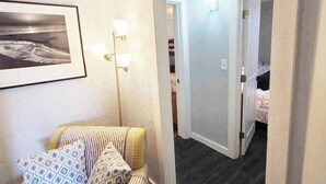 Une chambre d’hôtel avec un lit, une chaise, un tableau encadré au mur et un couloir menant à une autre pièce.