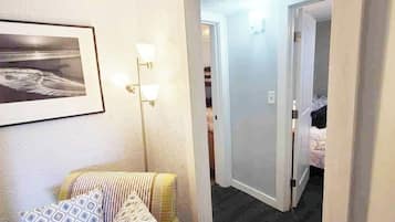 A hotel room with a bed, a chair, a framed picture on the wall, and a hallway leading to another room.