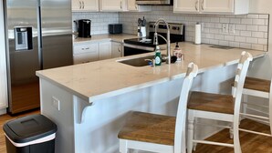 A modern kitchen with white cabinets, stainless steel appliances, a central island with bar stools, and a trash bin.