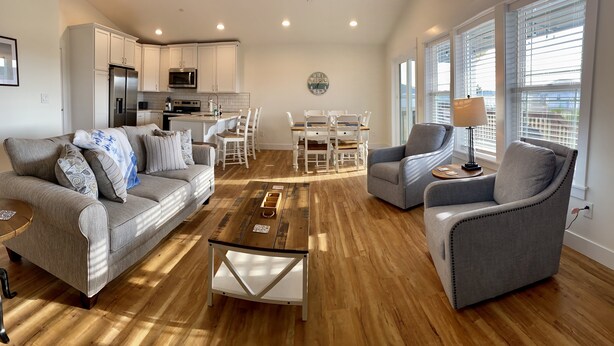 A spacious living area with a grey sofa, a coffee table, and two armchairs. A kitchen with white cabinets and a dining area with a table and chairs are visible in the background.