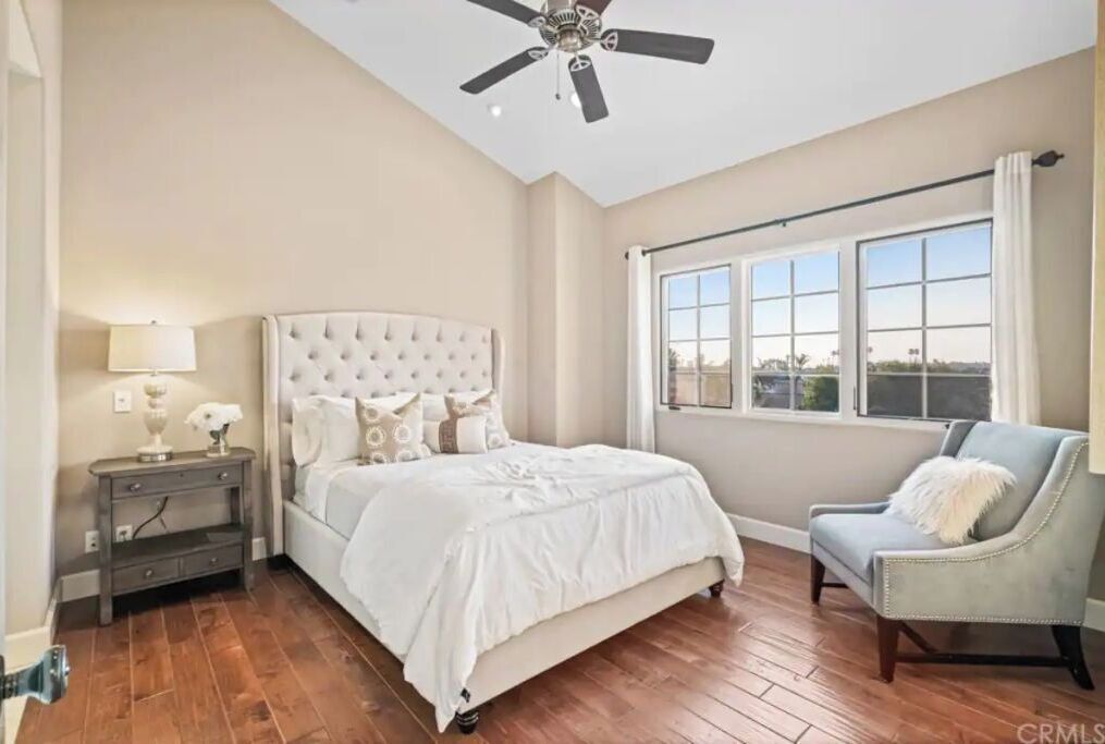 Une chambre à coucher avec un lit doté d’une tête de lit capitonnée, une table de chevet avec une lampe, une chaise et une grande fenêtre avec des rideaux.