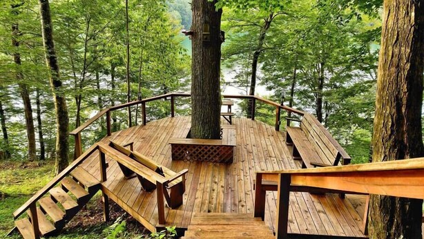 Een houten terras met banken en een open haard, omringd door bomen en een meer.