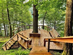 Eine Holzterrasse mit Bänken und Kamin, umgeben von Bäumen und einem See.