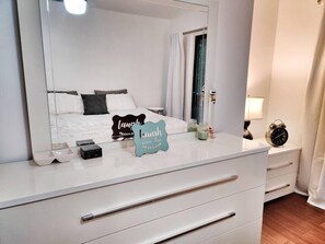 A bedroom with a large mirror, a bed with white and dark pillows, a bedside table with a lamp, a clock, and a sign that says "Laugh every day."