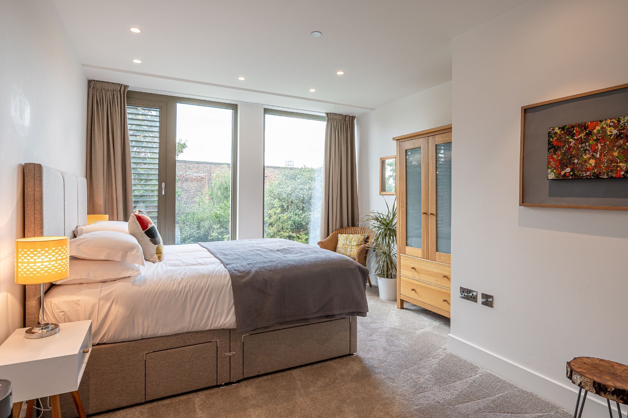 A modern bedroom with a large bed, a bedside table with a lamp, a wardrobe, and a painting on the wall.