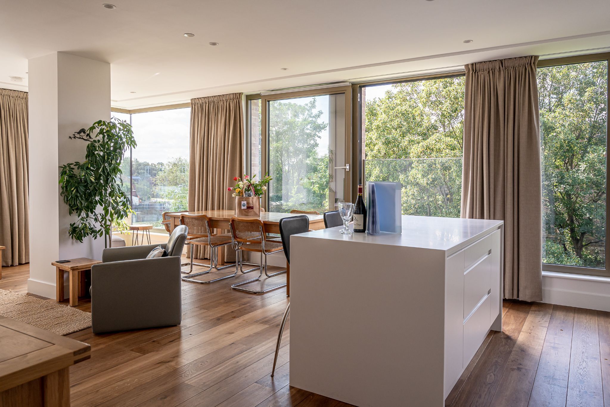 A modern living room with a wooden floor, a dining area with a table and chairs, a large window with curtains, and a potted plant.