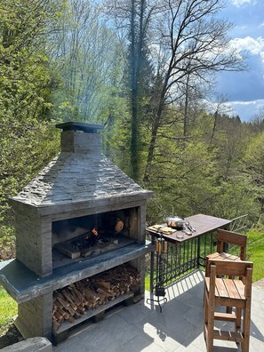 A stone-built outdoor fireplace with a fire burning, surrounded by a deck with a chair and a table. - Ideal oasis for a vacation surrounded with forest (Kupa, Brod na Kupi)