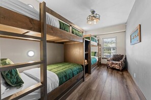 Een slaapkamer met stapelbed, een houten vloer, een enkel bed met groene beddengoed, een stoel met een kussen met luipaardprint en een plafondlamp.