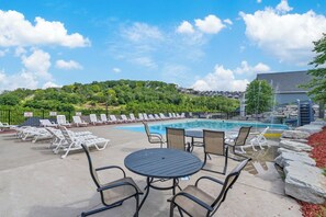 An outdoor pool area with lounge chairs, a table, and a view of a hill with trees and buildings. - Private Putting Green Retreat with Firepit, Hot Tub, Free Tickets, Views & Community Indoor Pool (Branson)