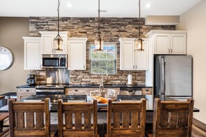 A kitchen with white cabinets, a stone backsplash, stainless steel appliances, and a dining area with wooden chairs. - Private Putting Green Retreat with Firepit, Hot Tub, Free Tickets, Views & Community Indoor Pool (Branson)