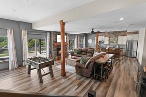 A spacious living area with a foosball table, a fireplace, a stone wall, and a kitchen in the background. - Private Putting Green Retreat with Firepit, Hot Tub, Free Tickets, Views & Community Indoor Pool (Branson)