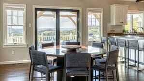 A dining area with a glass table surrounded by chairs, large windows with a view of hills, and a kitchen area with white cabinets and a bar. - 5BR Farmhouse☆King Size Bed☆Mountain View☆28 Acres (Ancramdale)