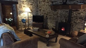 A cozy living room with a stone wall, a fireplace, a television, and a coffee table with apples. - Beautiful luxury restored gîte with private solar heated pool. (St. Paul la Roche)