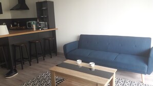 A compact living space with a sofa, a coffee table, a kitchen area with a microwave, and a bar stool. - Appartement salon cuisine chambre jardin. Sécurisé. Neuf. hygiène +++ (la Roche sur Foron)